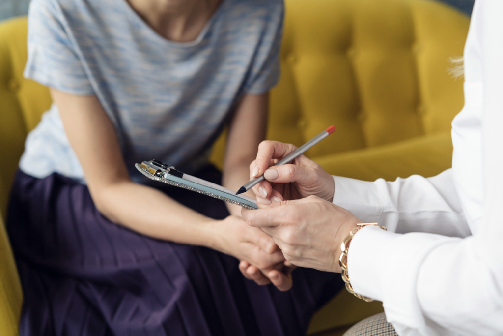 A clinician holds a clipboard while someone else sits on a couch.